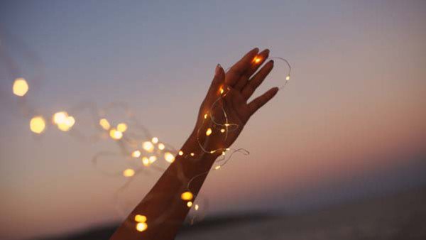 Female hand holding garland fairy string lighs against evening sky and sea. Romantic summer picture.