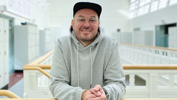 Dino står i Folkuniversitetets ljusgård. Han har svart keps, glasögon och en grå sweatshirt.