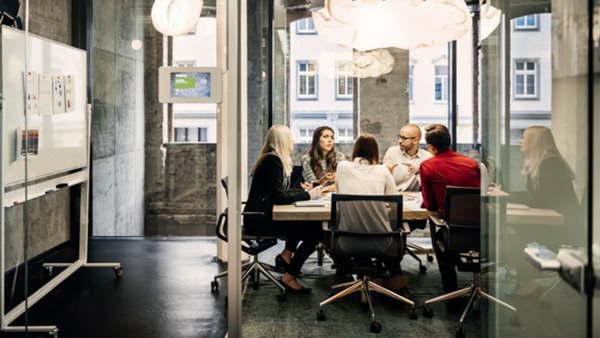 A group of people is sitting at a table during a business meeting in a bright, modern office. The team is talking business while pie charts can be seen hanging on the wall. Big bright windows are seen in the background.