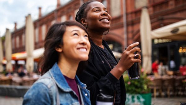 Smiling multi-ethnic female friends with drinks looking up while walking in city during vacation